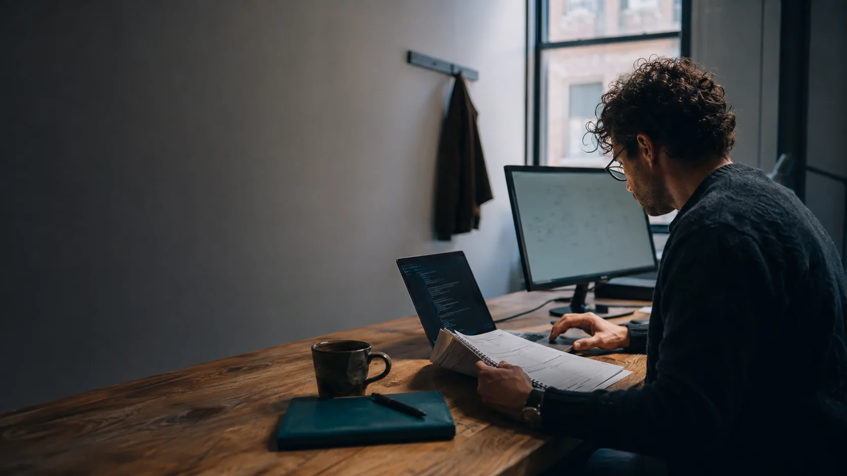 Une personne à une longue table de travail en bois lit un plan d'exécution imprimé et annoté au crayon tout en tapant sur un ordinateur portable, un second écran orienté de côté.
